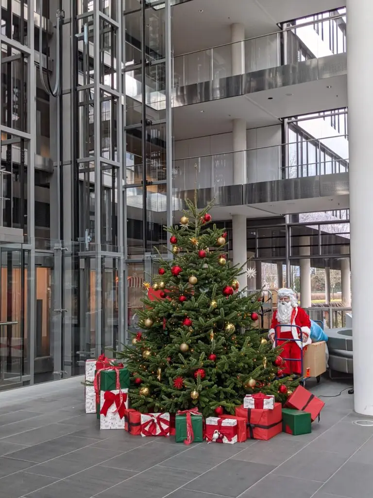 Foto einer großen Tanne, welche geschmückt ist mit einer Lichterkette und roten und goldenen Kugeln. Unter dem Baum liegen verpackte Geschenke in den Farben Grün, Weiß und Rot. Im Hintergrund der Tanne steh eine Weihnachtsmannpuppe.