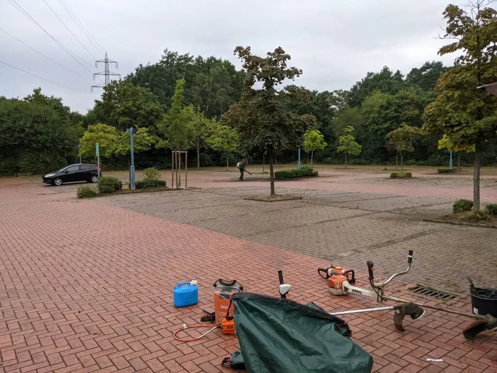 Foto von einer Parkplatzfläche und 2 Fadenschneidern. Im Hintergrund ist eine Person mit Rückengebläse.