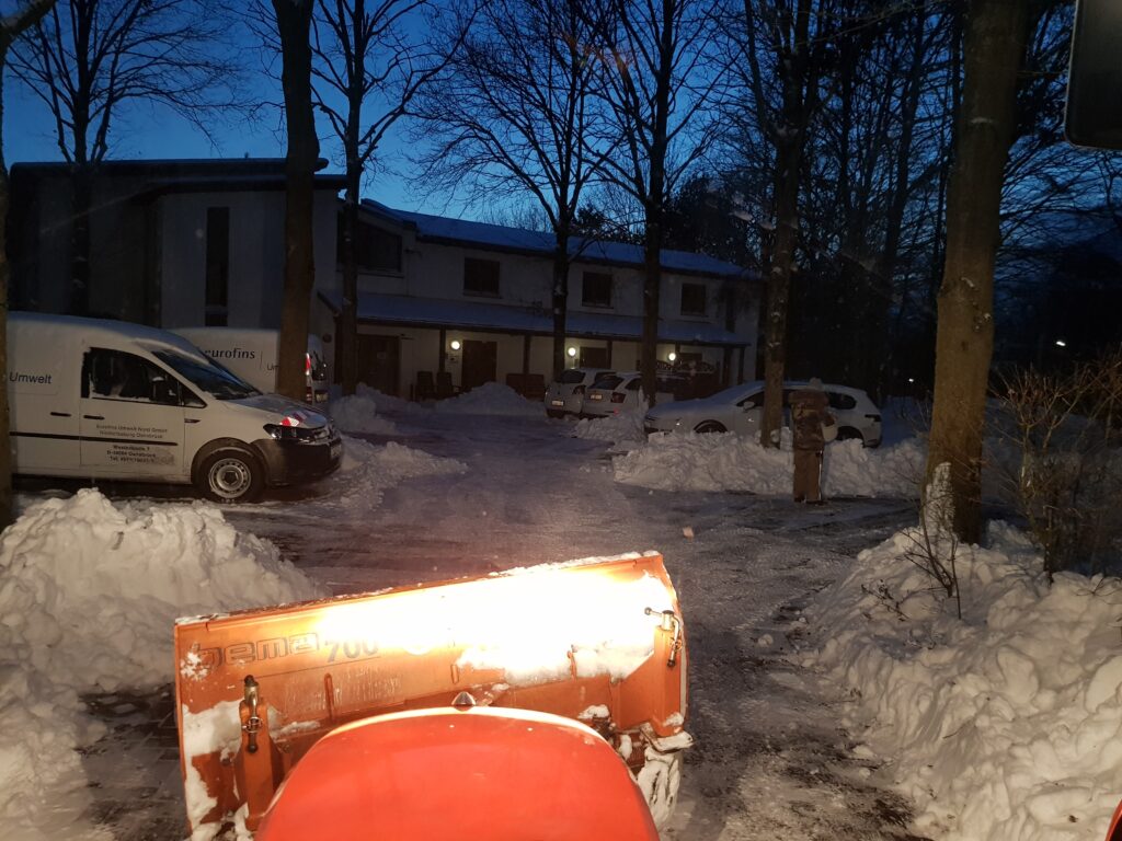 Foto aus Fahrerperspektive eines Schmalspurschleppers auf einen von Schnee frei geräumten Parkplatz bei Dunkelheit.