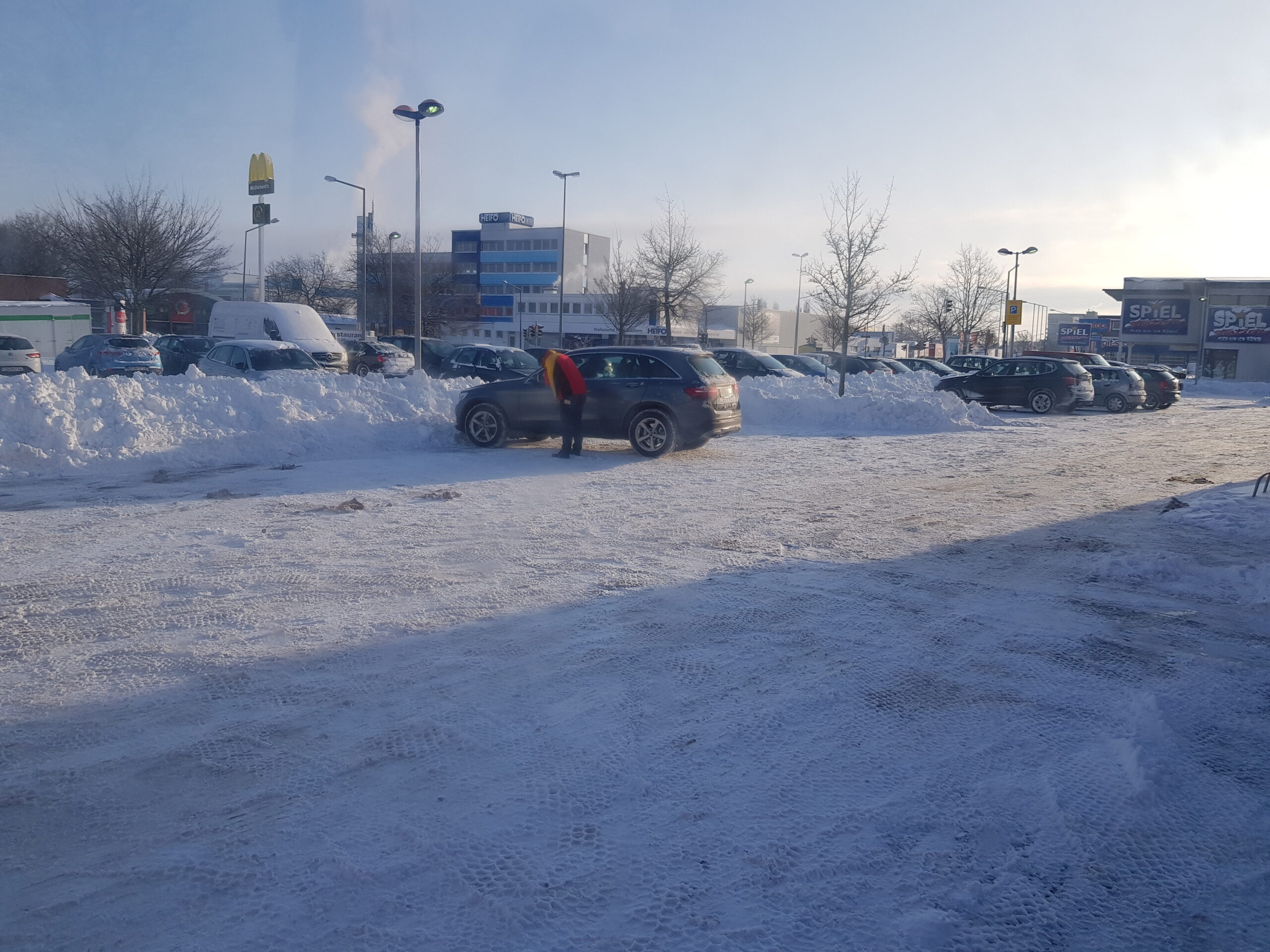 Foto von einem Parkplatz, der noch leicht mit Schnee bedeckt ist. Längliche Haufen mit Schnee liegen zwischen den parkenden Autos.