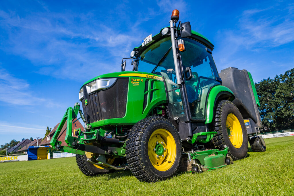 Foto von einem John Deere 3038R Schmalspurschlepper mit Mähwerk und Auffangkorb auf einem Sportplatz.