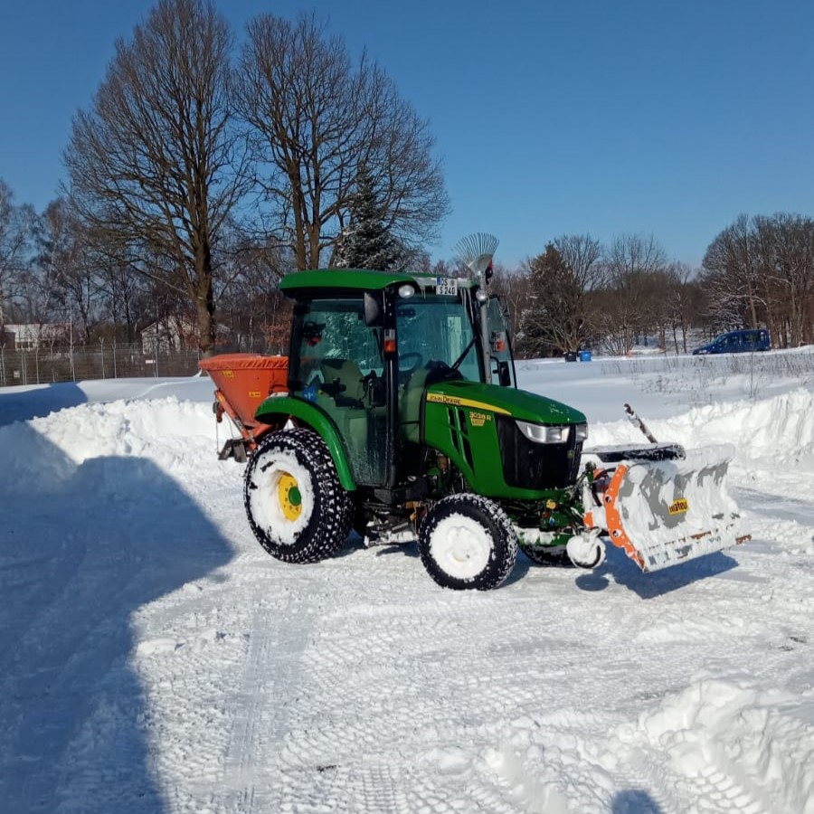 John Deere Schmalspurtrecker mit Schneeschild und Streuer in einer schneebedeckten Landschaft.